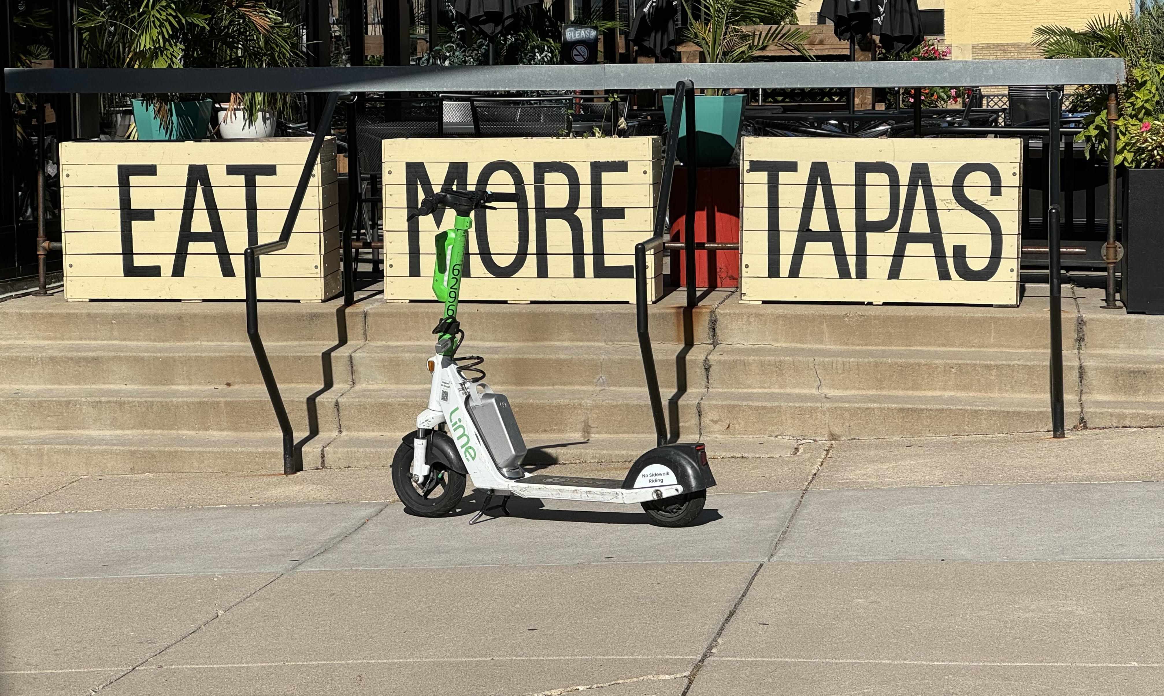 A green-and-white Lime scooter parked on a sunlit sidewalk in front of three cream wooden planters painted with the words EAT MORE TAPAS